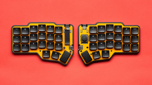 Fully assembled Unicorne LP keyboard in black with yellow plates, with Choc keycaps and OLEDs and joysticks, shown from top-down on red background